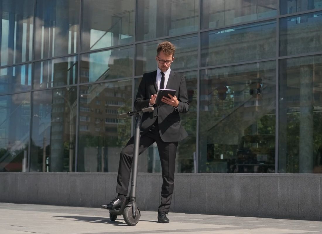 momak u odelu stoji na električnom trotinetu sa jednom nogom i gleda u tablet